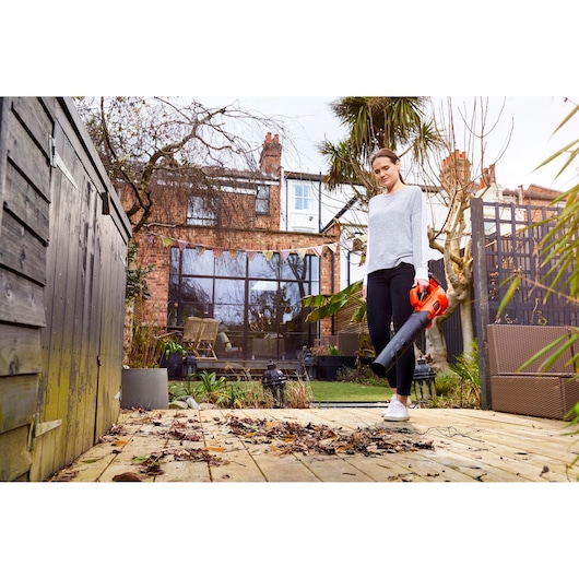 A person uses a BLACK+DECKER leaf blower (model BCBL200B) to clear leaves from a wooden deck in a residential garden, with a brick house and outdoor furniture in the background.