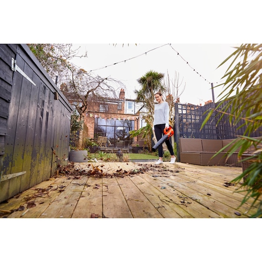 A person is using an orange BLACK+DECKER leaf blower to clear leaves and debris from a wooden deck in a garden. The scene features a residential house, plants, and garden furniture.