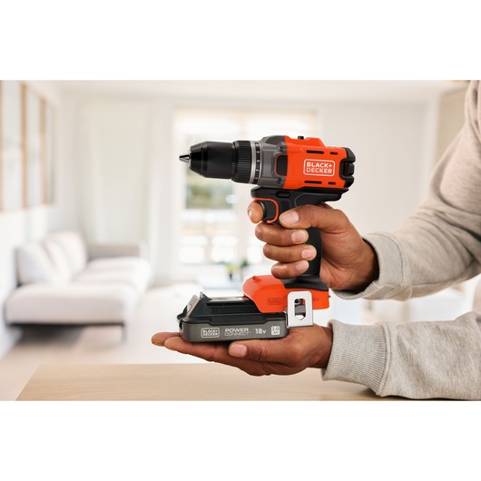 Person holding a BLACK+DECKER cordless drill inside a bright modern living room.