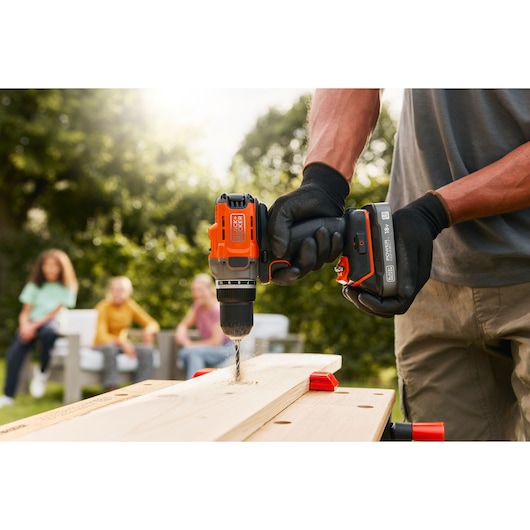 BLACK+DECKER BCD382D1 cordless drill in use, drilling into wood outdoors, with gloved hands holding the tool.