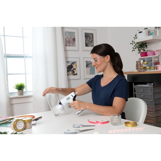 Photo of person using BLACKDECKER BCGL115 hot glue gun for crafts at a white table with jars and ribbons.