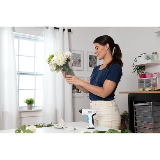 BLACK+DECKER BCGL115 cordless glue gun on table, woman arranging flowers in vase in bright crafting room.