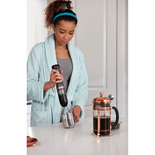 Photo of person using BLACK+DECKER BCKM1011KGF hand mixer with stainless steel frothing container in kitchen.