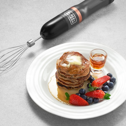 BLACK+DECKER BCKM101WHFF hand mixer with whisk attachment next to pancakes served with berries and syrup, top view.