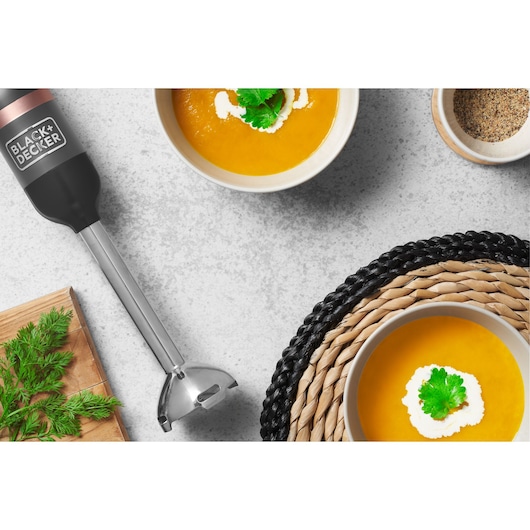 Photo of BLACK+DECKER hand blender BCKM1011KGF with stainless steel shaft beside bowls of soup and fresh herbs, top view.