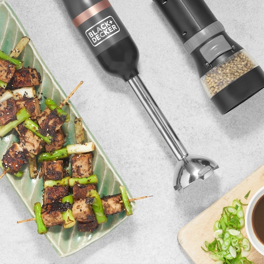 Photo of BLACK+DECKER BCKM1011KGF hand blender with stainless steel shaft, shown with food and spices on kitchen counter.