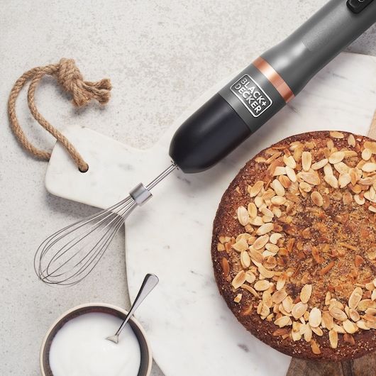 BLACK+DECKER BCKM101WHFF hand mixer with whisk attachment on marble board near almond cake and bowl of cream, top view.