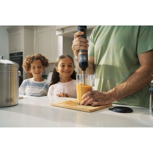 Photo of BLACK+DECKER BCKM1011KGF hand blender blending orange smoothie in glass, with kids watching in kitchen.