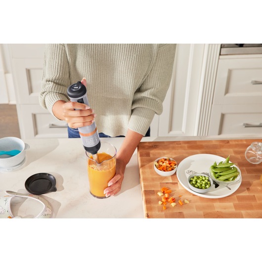 BLACK+DECKER BCKM1012KG hand blender mixing orange smoothie on kitchen counter next to chopped vegetables.