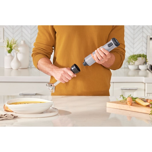 BLACKDECKER BCKM1012KG hand blender held in a kitchen, attachment being connected, soup and vegetables nearby.