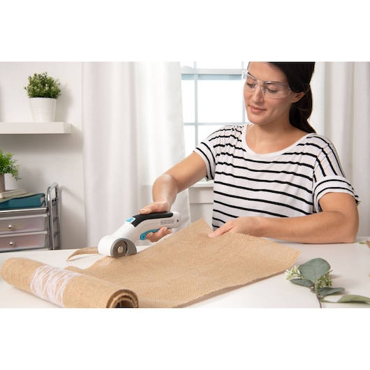 BLACKDECKER BCRC115 rotary cutter in use cutting burlap fabric on a tabletop in a bright room.