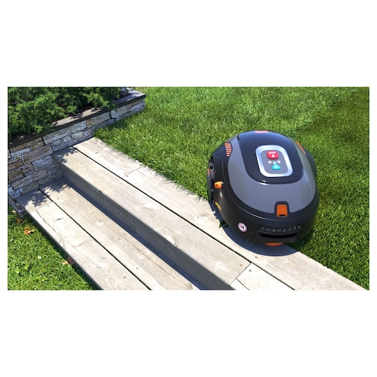A BLACK+DECKER robotic lawn mower is positioned on the edge of a grassy lawn next to a wooden pathway and stone wall.