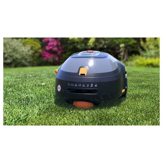 A BLACK+DECKER robotic lawn mower, model BCRMW122, operating on a green lawn with trees and flowers in the background.