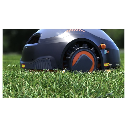 Close-up view of a BLACK+DECKER robotic lawn mower, model BCRMW122, on green grass outdoors. The mower has a modern design with black and orange detailing and visible wheels.