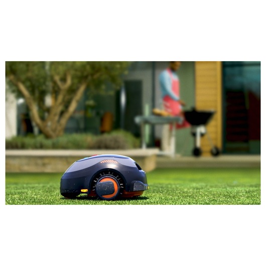 A BLACK+DECKER robotic lawn mower operates on a grassy lawn in a modern outdoor setting. In the background, a person stands near a barbecue grill, with trees and a house visible.