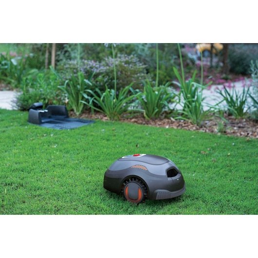 A BLACK+DECKER robotic lawn mower is operating on a grassy lawn, with its charging station visible in the background, surrounded by garden plants.