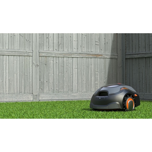 A robotic lawn mower on green grass in front of a wooden fence.