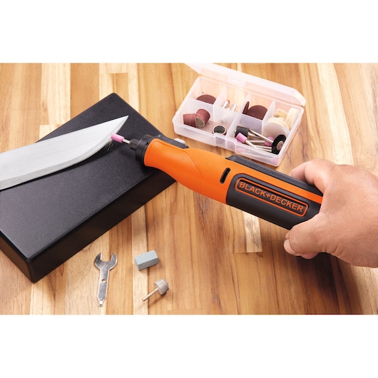 BLACK+DECKER BCRT8IK rotary tool in use sharpening knife, with accessory kit and attachments on wood table, side angle.
