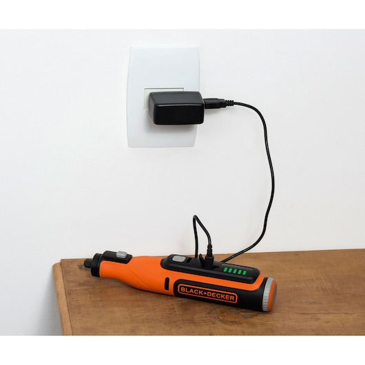 BLACK+DECKER BCRT8IK orange cordless rotary tool charging on a wooden table.
