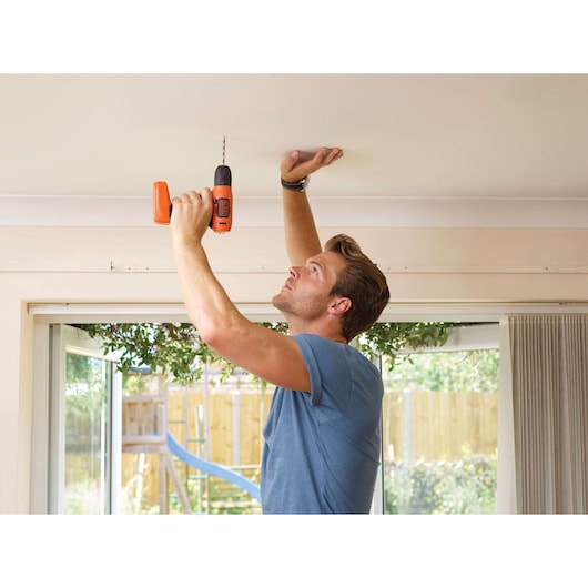 Person using BLACKDECKER BDCD8 cordless drill to drill into a ceiling indoors, garden visible through window.