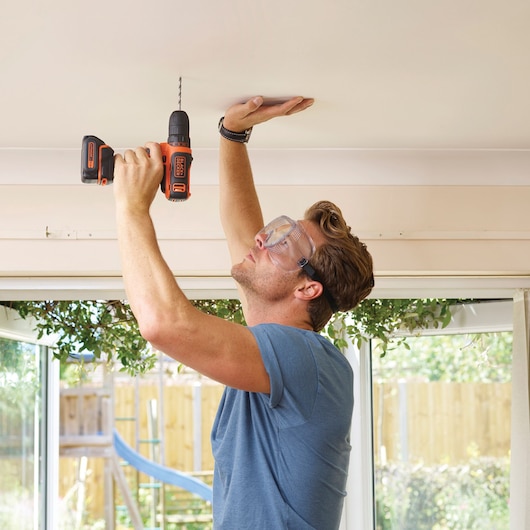 Photo of BLACKDECKER BDCDD121 cordless drill in use drilling into a ceiling indoors.