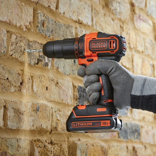 BLACKDECKER BDCHD18BS32 cordless drill in use, drilling into brick wall, lateral view, orange and black design.