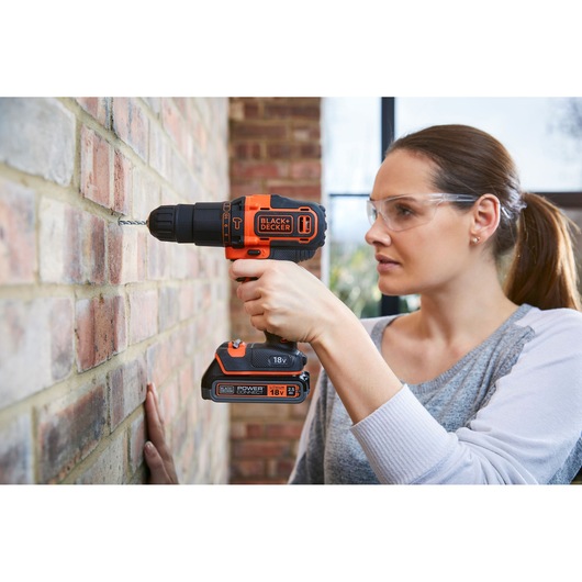 A person using a BLACK+DECKER cordless drill to drill into a brick wall. The drill is orange and black, and the person’s face is blurred for privacy.
