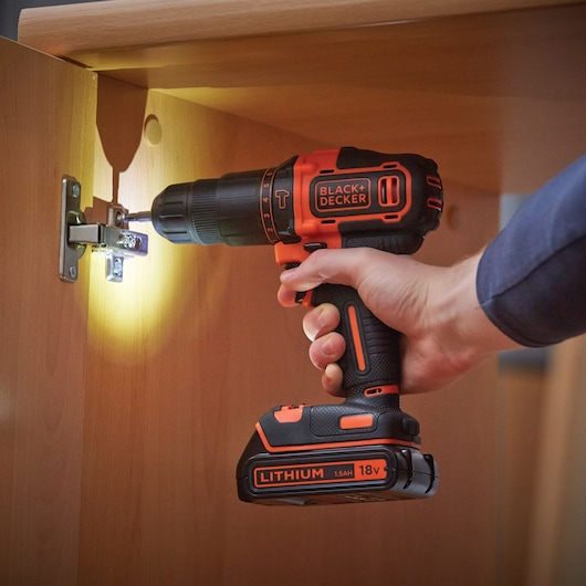 A person uses a BLACK+DECKER 18V cordless drill with lithium battery to screw a hinge on a wooden cabinet. The drill has an LED light illuminating the working area.