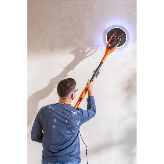Person uses a BLACK+DECKER electric drywall sander with LED light to smooth a wall surface.