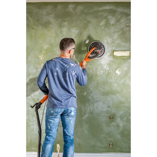 Person sanding a green wall with a BLACK+DECKER drywall sander, wearing goggles and work clothes.