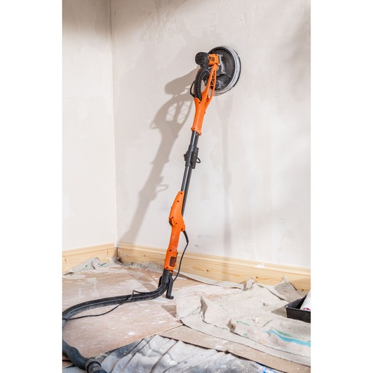 Orange drywall sander leaning against a wall, with a hose connected, used for smoothing plaster surfaces indoors.