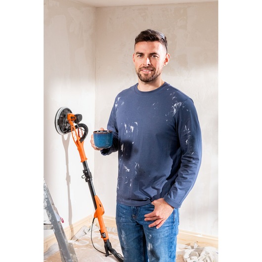 Man with paint-stained clothes holding mug, BLACK+DECKER wall sander and ladder beside him in a room under renovation.