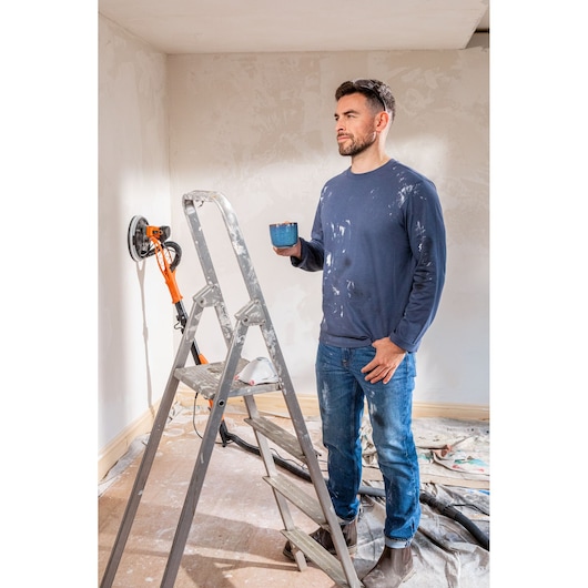 A person in work clothes stands near a ladder and a BLACK+DECKER wall sander, holding a mug during a room renovation.