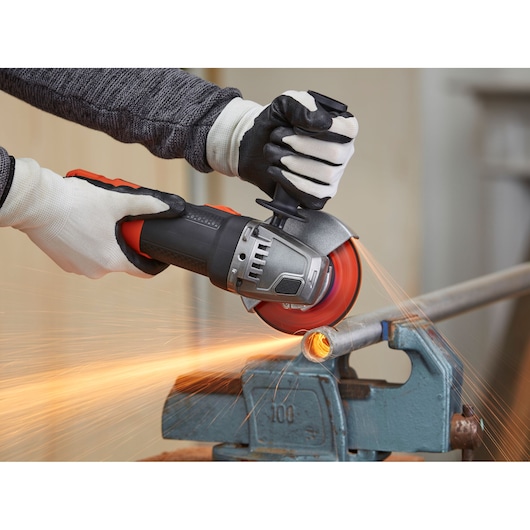 A person wearing protective gloves uses a BLACK+DECKER angle grinder to cut through a metal pipe, creating sparks.