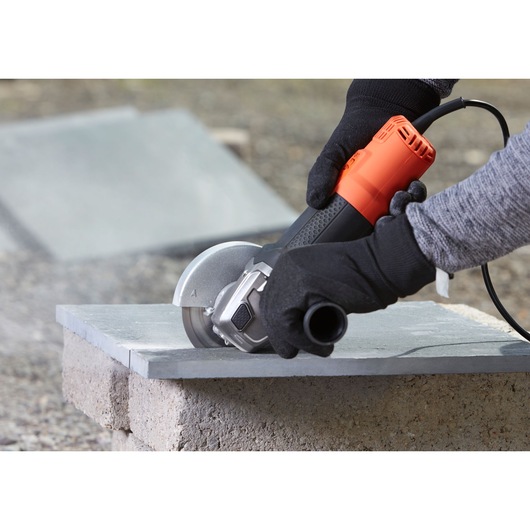 A person wearing black gloves uses a BLACK+DECKER BEG210 angle grinder to cut a stone tile outdoors.