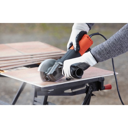 Person wearing gloves using a BLACK+DECKER BEG210 angle grinder to cut a tile on a workbench outdoors.