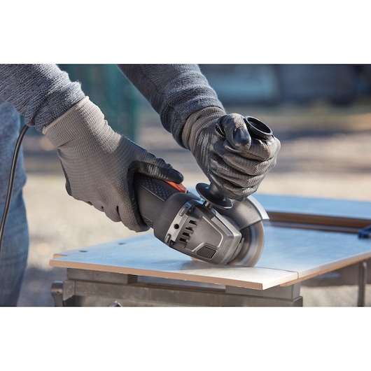 Close-up of gloved hands operating a BLACK+DECKER BEG210 angle grinder to cut a tile on a workbench outdoors.