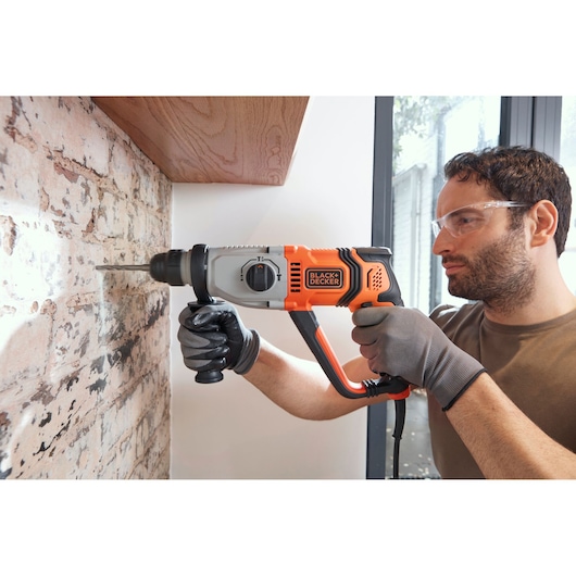 BLACK+DECKER BEHS02K hammer drill in use on brick wall, orange and black design, person wearing gloves.