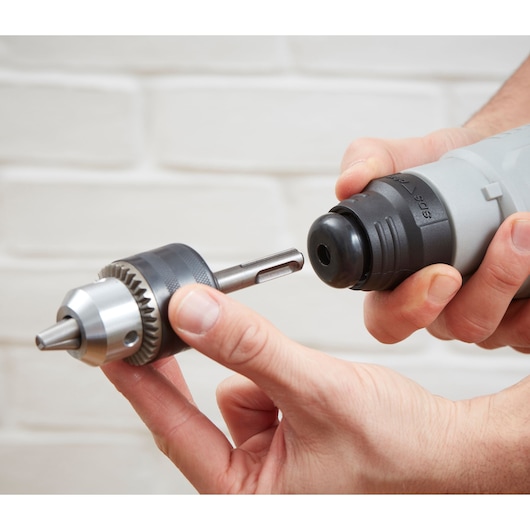 Close-up photo of BLACKDECKER BEHS02K drill with keyless chuck attachment held by hands against a white brick wall.