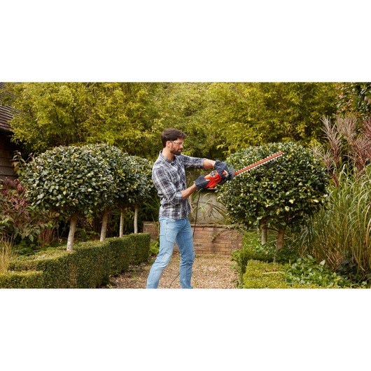 BLACKDECKER BEHT251KIT hedge trimmer used in garden, red and black tool cutting shrubbery outdoors.