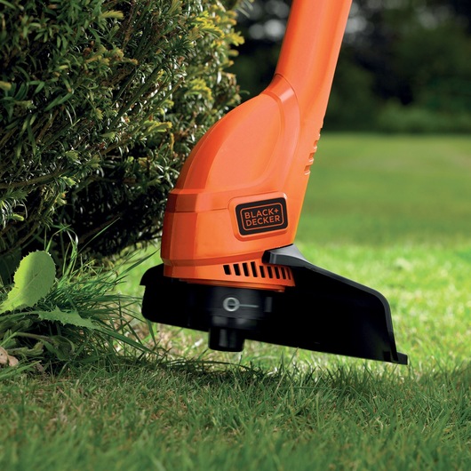 BLACK+DECKER BEMW481BGL2 orange grass trimmer cutting grass near a bush in a garden.