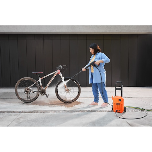 BLACK+DECKER BEPW1300L pressure washer cleaning a muddy bicycle outdoors, person holding spray wand.