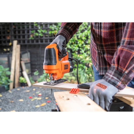 Photo of BLACKDECKER BES602 jigsaw in use, cutting wood outdoors, person wearing gloves and plaid shirt.