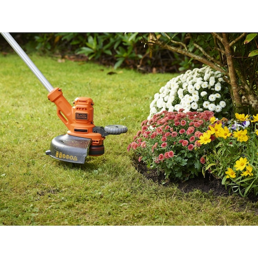 BLACKDECKER BESTA530C10 orange grass trimmer in use trimming lawn edge near flowering plants.