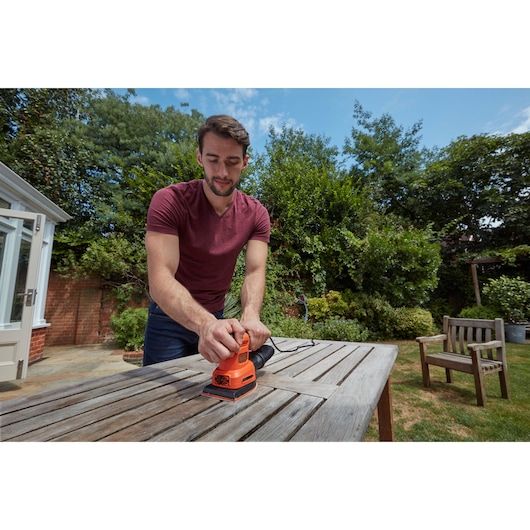 BLACKDECKER BEW200 sander in use on outdoor wooden table, orange tool held by person in garden setting.