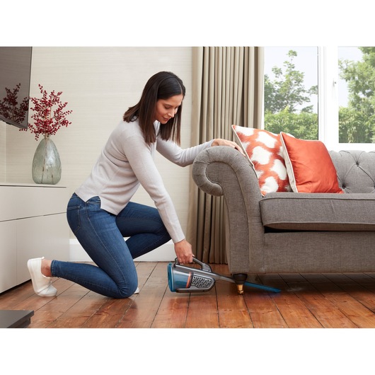 BLACKDECKER BHHV320J handheld vacuum cleaning under gray sofa in living room with orange pillows.