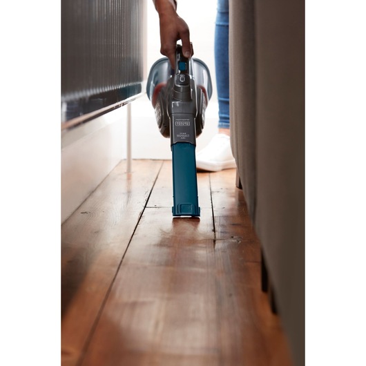 BLACKDECKER BHHV320J handheld vacuum cleaning narrow space beside furniture on wooden floor.