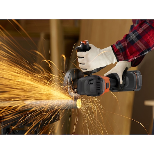 A person wearing protective gloves uses a BLACK+DECKER cordless angle grinder to cut through a metal pipe, creating bright, flying sparks.