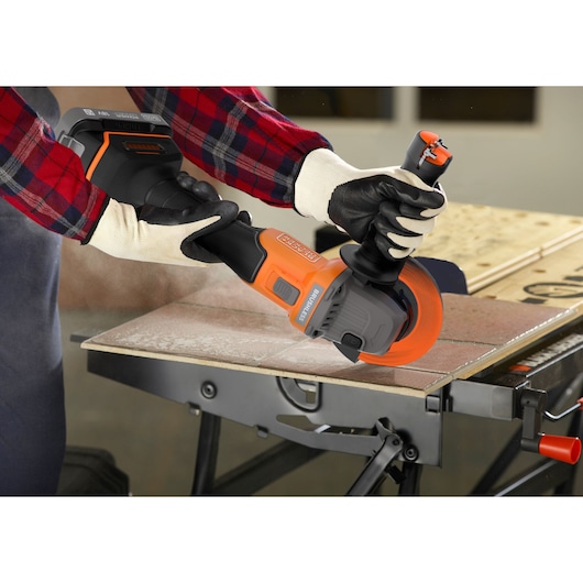 A person wearing gloves uses a BLACK+DECKER cordless angle grinder to cut a tile on a workbench. The tool has an orange and black design and is creating dust during the cutting process.