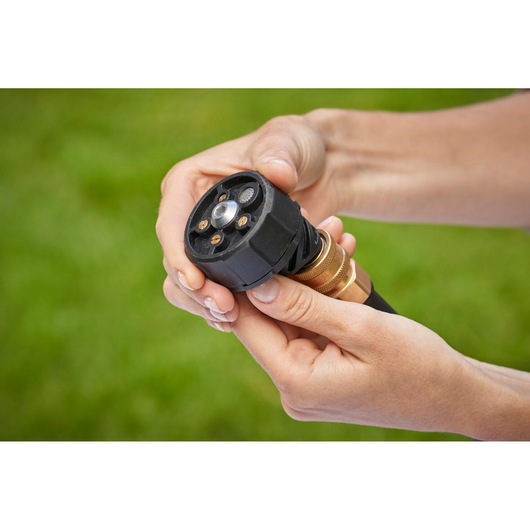 Close-up of hands adjusting a Black+Decker garden hose nozzle with multiple spray settings, shown against a blurred green background.